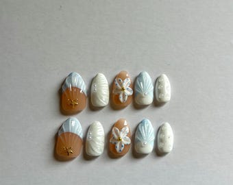 May include: A set of ten almond-shaped press-on nails featuring a beach-inspired design. The nails include light blue and white shell patterns, raised white floral accents with gold centers, small gold starfish charms, and white pearl embellishments.