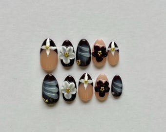 May include: A set of ten oval-shaped press-on nails featuring a dark brown and nude color palette. The designs include white and dark brown floral embellishments with gold centers, along with subtle white brushstroke patterns on dark brown bases.