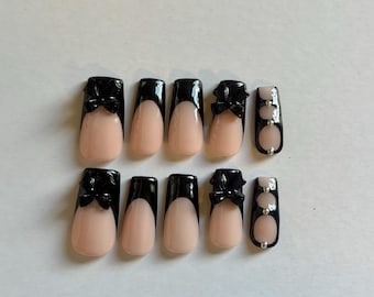 May include: A set of ten press-on nails featuring a nude base with black French tips. Some nails are adorned with three-dimensional black bows, while others feature a vertical black stripe accented with small silver metallic studs.