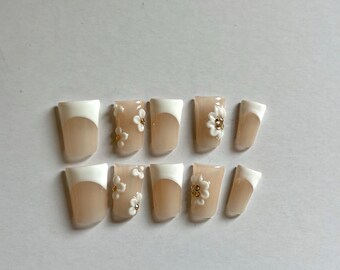 May include: A set of ten press-on nails featuring a nude base with white French tips. Several nails are decorated with three-dimensional white flower appliqués that have small gold-colored centers, arranged in a square-shaped nail design.