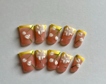May include: A set of ten press-on nails featuring a peach-colored base with yellow French tips. Each nail is decorated with three-dimensional white floral accents, green stems, and small silver-toned studs arranged in a floral pattern.