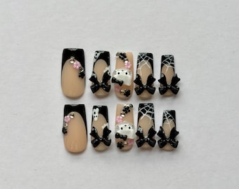 May include: A set of ten press-on nails featuring a nude base with black tips, white spiderweb patterns, and three-dimensional embellishments including black bows, small pink flowers, and white cat-shaped charms with black details.