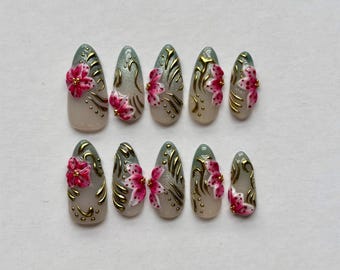May include: A set of ten press-on nails featuring a gradient base transitioning from a soft beige to a muted sage green. Each nail is decorated with raised pink floral accents, gold metallic swirls, and small gold dots across the surface.