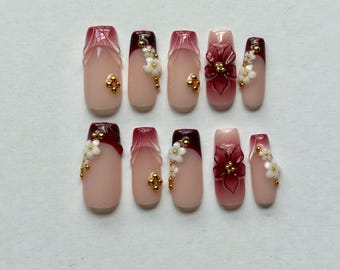 May include: A set of ten press-on nails featuring a nude base with deep burgundy French tips and gradient designs. The nails are decorated with three-dimensional white floral accents and small gold metallic beads arranged in clusters.