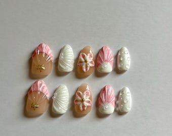 May include: A set of ten press-on nails featuring a variety of designs including pink and white shell patterns, raised white floral accents with gold centers, small gold starfish charms, and white nails with raised circular dot textures.