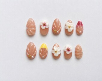 May include: A set of ten press-on nails in a nude base color, featuring raised white floral accents, gold-toned starfish charms, small white pearls, and textured shell-like ridges. Some nails include yellow or pink tips with a glossy finish.