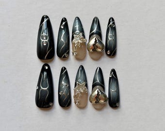 May include: A set of ten almond-shaped press-on nails featuring a black and gray gradient finish with silver line accents and small rhinestones. Two nails feature a silver star charm, and two nails feature a silver broken heart charm.