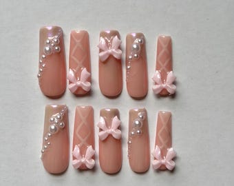 May include: A set of ten press-on nails in a soft peach tone with a glossy finish. The design features raised light pink bows, white pearl accents arranged in a curved line, and a subtle crisscross pattern embossed on the surface of the nails.