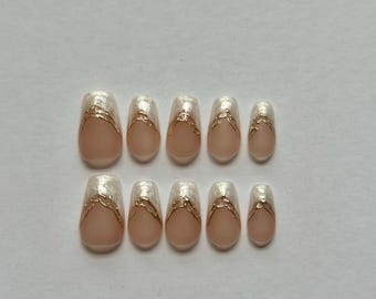 May include: A set of ten press-on nails featuring a nude base with a pearlescent white tip. Each nail is accented with a thin, metallic gold line that creates a V-shaped design across the center, separating the nude and pearlescent sections.