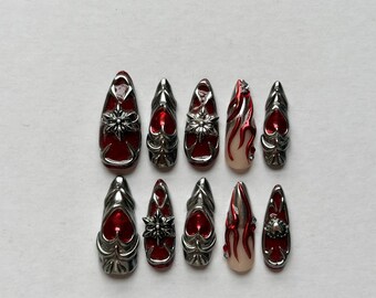 May include: A set of ten press-on nails featuring a pointed almond shape. The designs include deep red glossy bases with metallic silver 3D embellishments, including star-like charms, heart accents, and raised flame patterns on a nude base.