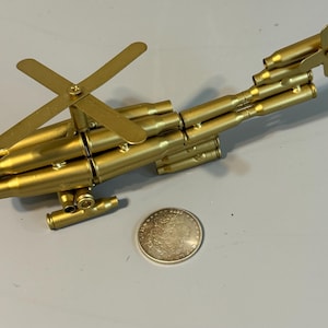 May include: A handcrafted, gold-colored metal helicopter sculpture. The model is constructed from bullet casings and features a rotor and tail rotor. A silver coin is placed next to the sculpture for scale.