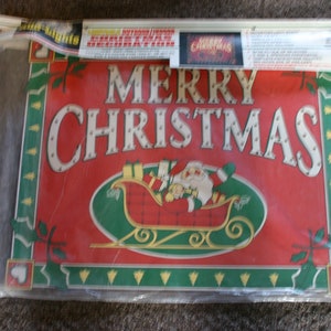 May include: A red and green Christmas sign with the words "Merry Christmas" in white letters with white dots. A cartoon Santa Claus is in a sleigh with presents.