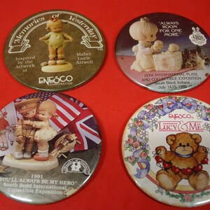 May include: Four round pinback buttons featuring Precious Moments figurines. The top left button features a girl in a green dress with the text "Memories of Yesterday". The top right button features a girl holding a baby with the text "Always Room for One More". The bottom left button features a girl and boy in a patriotic scene with the text "You'll Always Be My Hero". The bottom right button features a teddy bear with the text "Lucy & Me".