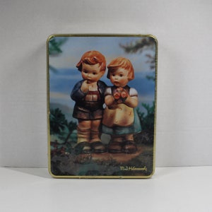 May include: A vintage tin box with a colorful image of two children. The boy is wearing a blue shirt and brown pants. The girl is wearing a green dress with a yellow apron. The tin box has a gold trim and the text "M.J. Hummel" on the bottom.