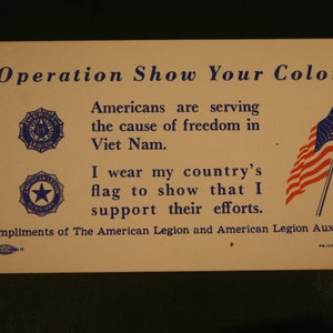 May include: A white card with blue text that reads "Operation Show Your Colors". The text continues, "Americans are serving the cause of freedom in Viet Nam. I wear my country's flag to show that I support their efforts." The card also features a blue and white American Legion logo and a small American flag in the upper right corner. The text at the bottom reads "Compliments of The American Legion and American Legion Auxiliary."