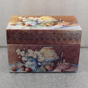 May include: A rectangular box with a hinged lid, featuring a vintage still-life design. The artwork depicts a blue vase with white flowers, a lamp, and fruit. The box has a brown background with a decorative floral border.