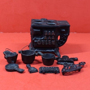 May include: A miniature black cast iron stove with the word QUEEN embossed on the front, accompanied by a collection of small kitchen accessories including two handled pots, a skillet, a shovel, a rectangular grate, and a decorative iron stand.