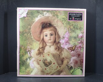 May include: A vintage-style porcelain doll wearing a pink hat and dress, holding a lace-trimmed card with a floral design. The doll is surrounded by pink flowers and a butterfly. The box reads "Jigsaw Puzzle Over 500 Pieces by Cobblestone".