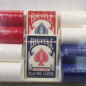 Pu&ograve; includere: Un set confezionato di forniture da poker, tra cui due mazzi di carte da gioco Bicycle Rider Back e pile di fiches da poker rosse, bianche e blu. Le carte sono in una scatola bianca con dettagli rossi e blu.