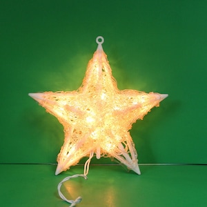 May include: A glowing, star-shaped Christmas decoration. The star is made of white plastic and has a woven design. It is lit with warm white lights and has a hanging loop at the top. The star is on a green background.