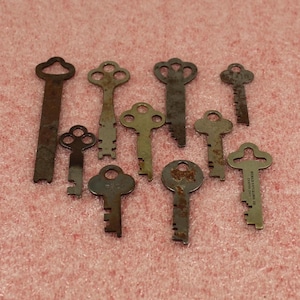 May include: A collection of antique keys in various shapes and sizes, arranged on a pink surface. The keys are made of metal and show signs of age, with rust and wear visible. Some keys have ornate designs, while others are simple.