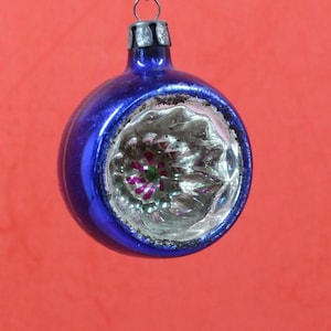 May include: A vintage Christmas ornament with a deep blue exterior and a silver, textured interior. The ornament has a circular shape with a small metal cap and a loop for hanging. The interior features a floral design with pink and green accents.