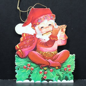 May include: A red and white Christmas ornament featuring a cartoon elf playing a flute while sitting on a bed of green holly with red berries.