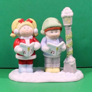 May include: A ceramic Christmas figurine featuring two children singing carols from a book labeled "NOEL". One child wears a red outfit, the other a blue coat and green hat. A snow-covered base and a lamppost complete the scene.