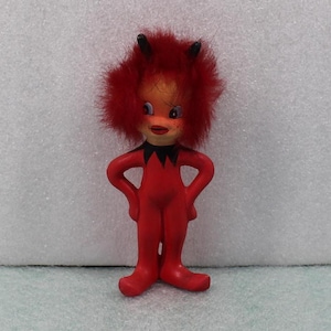 May include: A vintage devil figurine with a red body, red fur hair, and black horns. The figurine has a smiling face with blue eyes and is posed with hands on hips. The devil is wearing a black collar.