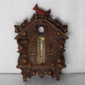 May include: A decorative wooden thermometer shaped like a house, with a red bird perched on top. The thermometer displays temperature readings in Fahrenheit. The house is intricately carved with floral and architectural details.