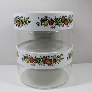 May include: A set of two clear glass canisters with white lids and a floral pattern featuring vegetables and herbs. The lids have the text "Fire King Made in U.S.A. Better Homes and Gardens" printed on them.