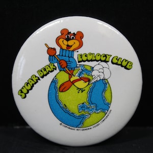 May include: White button with a cartoon bear character wearing a blue sweater and holding a broom, standing on a globe. The text reads "Sugar Bear Ecology Club" in green and yellow. Copyright 1971 General Foods Corporation.