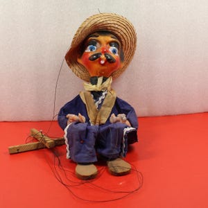 May include: A vintage marionette doll featuring a painted face with a mustache and straw hat. The doll wears a blue outfit with a tan shawl and white trim. The puppet has wooden feet and hands, with strings attached for manipulation.