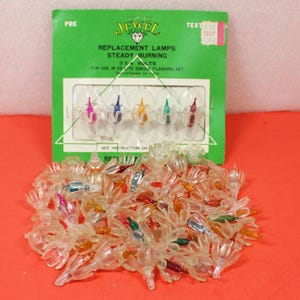 May include: A package of Jewel replacement lamps for a 35-light flashing set. The green packaging displays the text "REPLACEMENT LAMPS STEADY BURNING 3.54 VOLTS". The package contains five multi-colored bulbs, with a pile of clear plastic bulb holders in front.