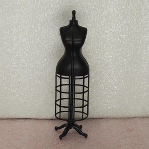May include: A matte black miniature dress form mannequin featuring a solid torso and a cage-style skirt structure. The display piece stands on a four-legged base and includes a small knob at the neck, providing a structured frame for clothing.