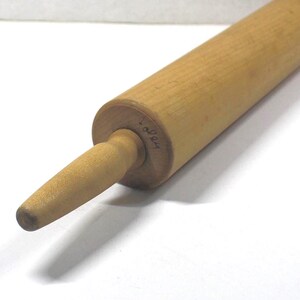 May include: A light brown wooden rolling pin with a cylindrical body and a tapered handle. The end of the cylinder features a small, handwritten inscription that appears to read 1024 in dark ink, visible on the wood grain surface.