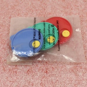 May include: Three colorful Tupperware Crystalwave seal stack magnets in a clear plastic bag. The magnets are blue, green, and red with yellow accents. The bag has text that reads "CRYSTALWAVE SEAL STACK MAGNET" and a warning about small parts.