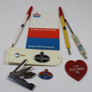 May include: A collection of vintage Amoco brand promotional items, including a pen, a notepad, a multi-tool keychain, a patch, a screwdriver, and a heart-shaped keychain with the text "I Love Atlas/Amoco TBA".