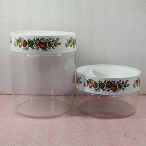 May include: Two clear, cylindrical storage containers with white lids. The lids feature a decorative band with colorful illustrations of fruits and vegetables. One container is taller than the other, suggesting different sizes for various storage needs.