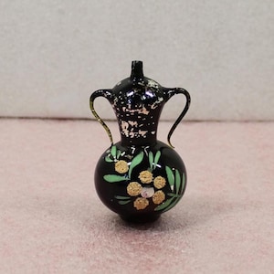 May include: A small, black decorative vase with a round base and two handles. The vase features gold and green floral designs. The top of the vase has a lid and a small spout. The vase is approximately 4 inches tall.