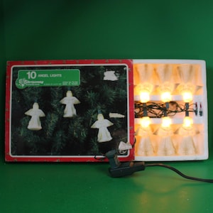May include: A box of 10 white angel Christmas lights with a green wire. The box has a red border and says "10 Angel Lights" on the front. The lights are in a white styrofoam box with a clear plastic cover. The lights are plugged in and lit.