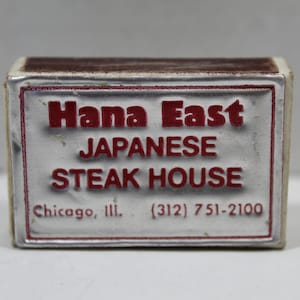 May include: A silver matchbook with red lettering that reads "Hana East Japanese Steak House" and "Chicago, Ill. (312) 751-2100".