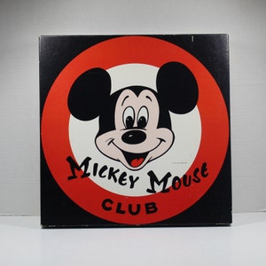 May include: A black and red circular sign with the words "Mickey Mouse Club" in white lettering. The center of the sign features a cartoon image of Mickey Mouse with a white background.