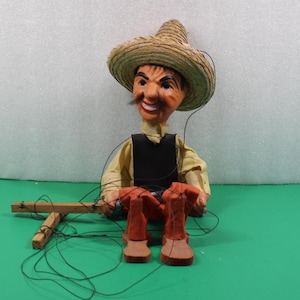 May include: A vintage marionette doll of a man wearing a straw hat, yellow shirt, black vest, and orange pants. The doll has a painted face with a mustache and is controlled by strings attached to a wooden crossbar. The doll is sitting.