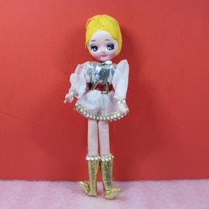 May include: A vintage doll with a yellow yarn wig, painted facial features, and a white dress with silver sequin bodice and red belt. The doll has gold sequined boots and is posed against a red background.