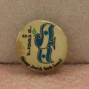 May include: A round pinback button with a cream-colored background. It features a blue graphic of a dove carrying a green olive branch, with two blue Stars of David. Text reads 22, Hebrew characters, and Chicago Jewish Youth Council.
