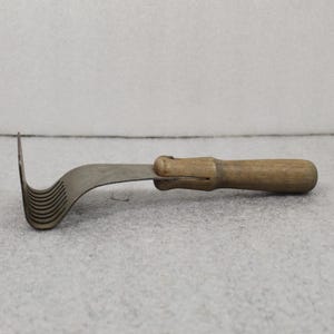 May include: An antique kitchen tool with a wooden handle and a curved, metal blade. The blade has a series of parallel, curved slots. The handle is a light brown color, and the metal blade has a dull, silver finish. The tool appears to be used for slicing or cutting.