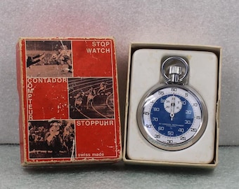 Puede incluir: Un cronómetro vintage con esfera azul y carcasa plateada, presentado en su caja roja original. La caja presenta ilustraciones de atletas y las palabras "STOP WATCH", "CONTADOR" y "STOPPUHR". El cronómetro está marcado "swiss made".