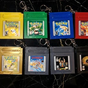 May include: A collection of eight Game Boy cartridge keychains in various colors. The keychains feature game titles like Pokémon, Super Mario Land, and WWF. Each cartridge has a silver keyring attached.