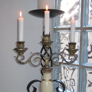 Puede incluir: Un candelabro de tres brazos con tres velas encendidas. El candelabro está hecho de metal con un acabado dorado y tiene una base de cerámica blanca. El candelabro está sobre una superficie de mármol.